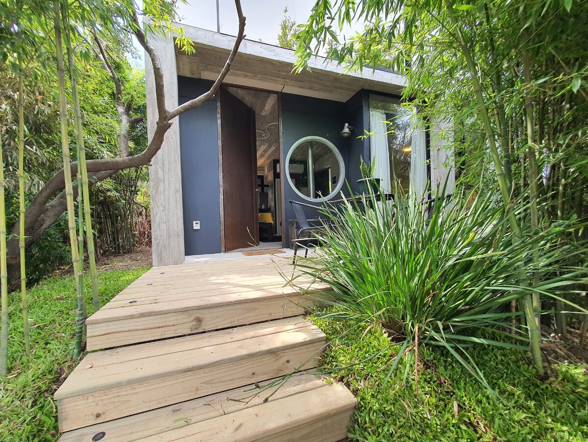 A modern structure framed by lush greenery is approached via wooden steps. The entrance features a circular window that offers a glimpse into the serene interior. Bamboo and vibrant plants surround the space, creating a natural setting.