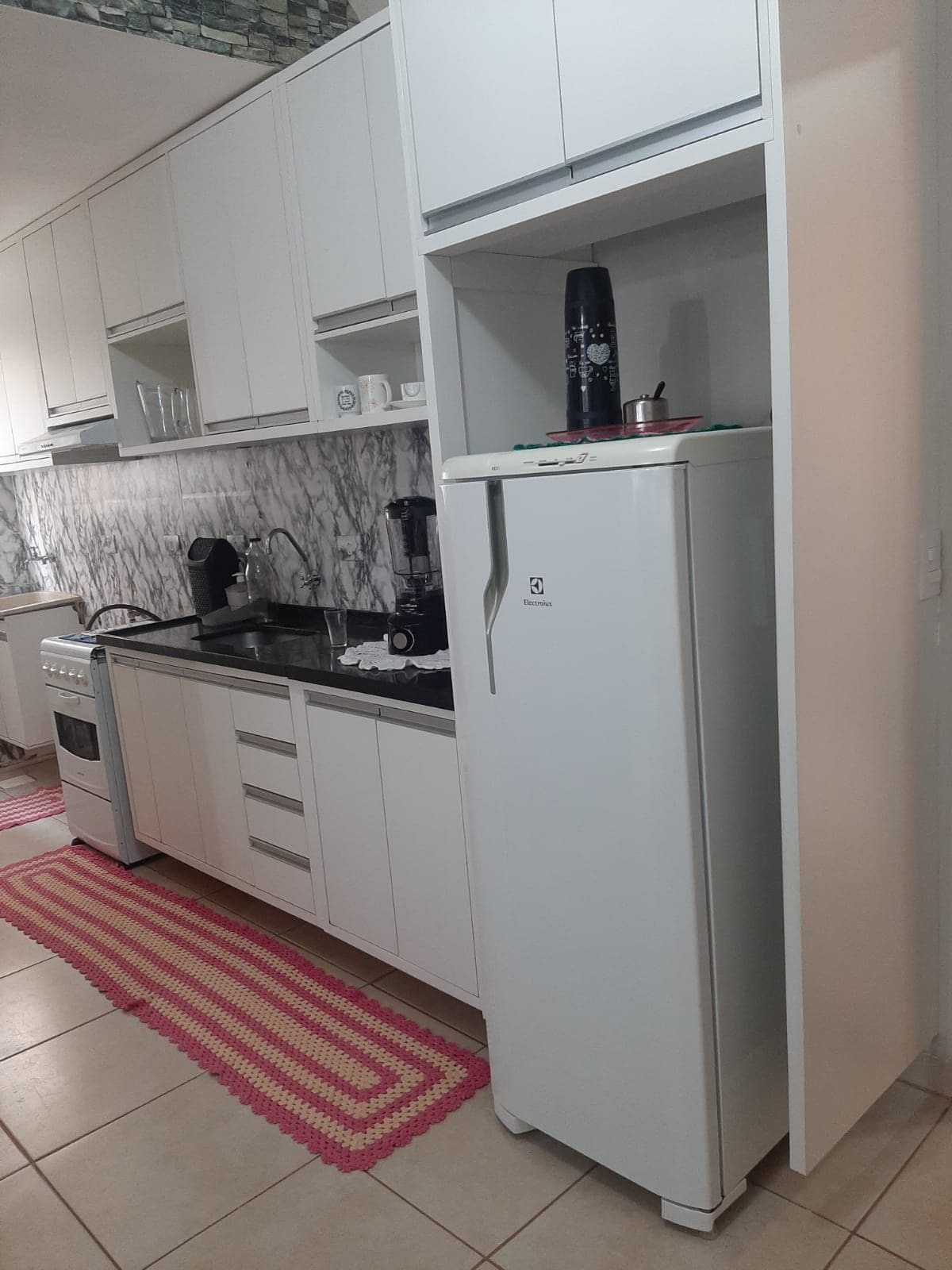 A modern kitchen showcases sleek white cabinetry and dark countertops. A refrigerator is positioned alongside a stovetop and a kitchen sink, with a marble backsplash adding texture. A rug in warm tones lines the floor, providing a contrasting detail to the overall design.