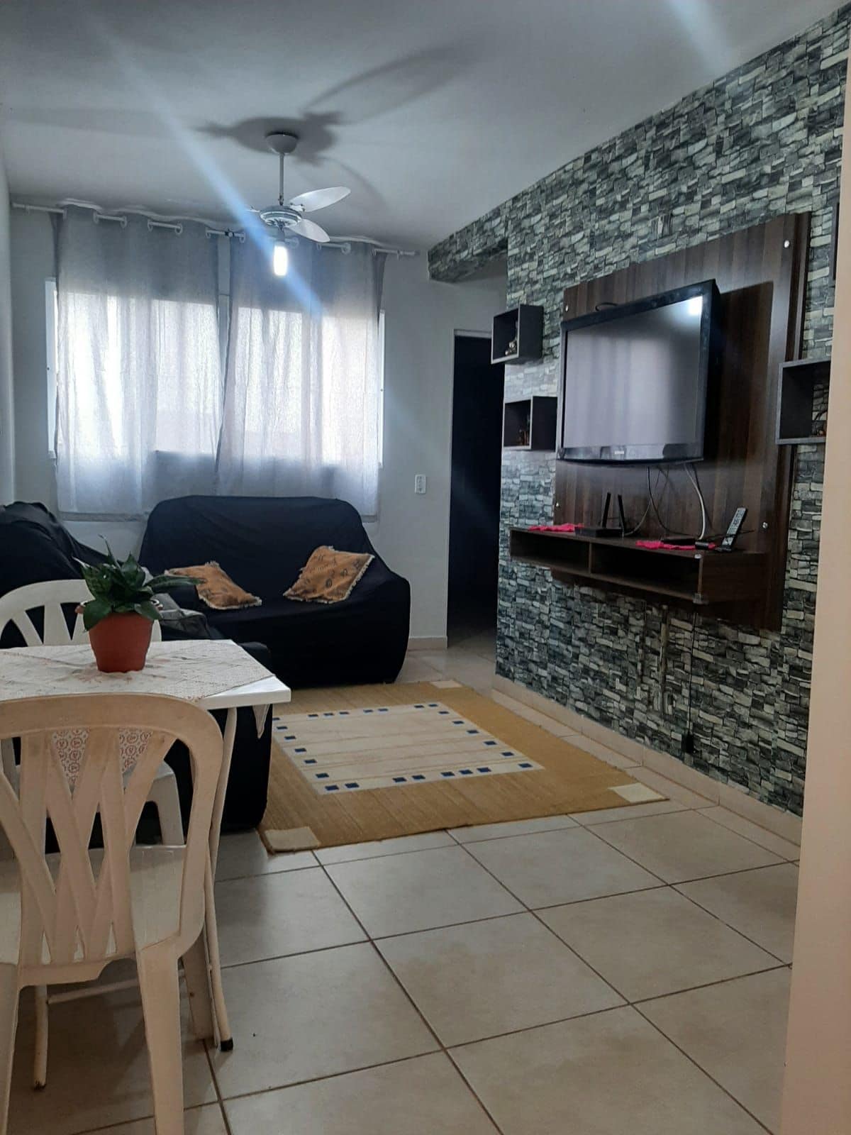 A welcoming living area is presented, featuring a ceiling fan and natural light filtering through sheer curtains. A small dining table is positioned beside a cozy seating area, with a television mounted on a stylish stone wall. A patterned rug adds warmth to the space.