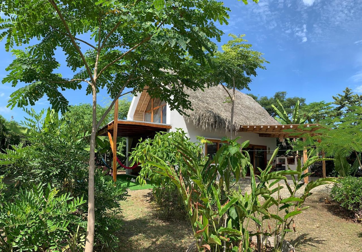 A two-story house with a thatched roof is surrounded by lush greenery. Large glass doors lead to a covered outdoor area, offering a smooth transition between nature and living space. Tropical plants and trees enhance the outdoor setting, contributing to a serene atmosphere.
