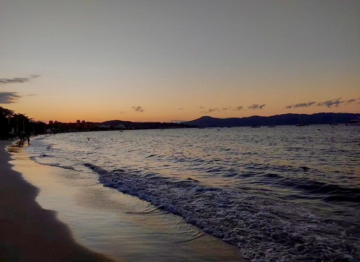The image captures a serene beach scene at sunset, with gentle waves lapping at the shore. Soft hues of orange and pink fill the sky, reflecting on the water, while silhouettes of distant mountains provide a tranquil backdrop.