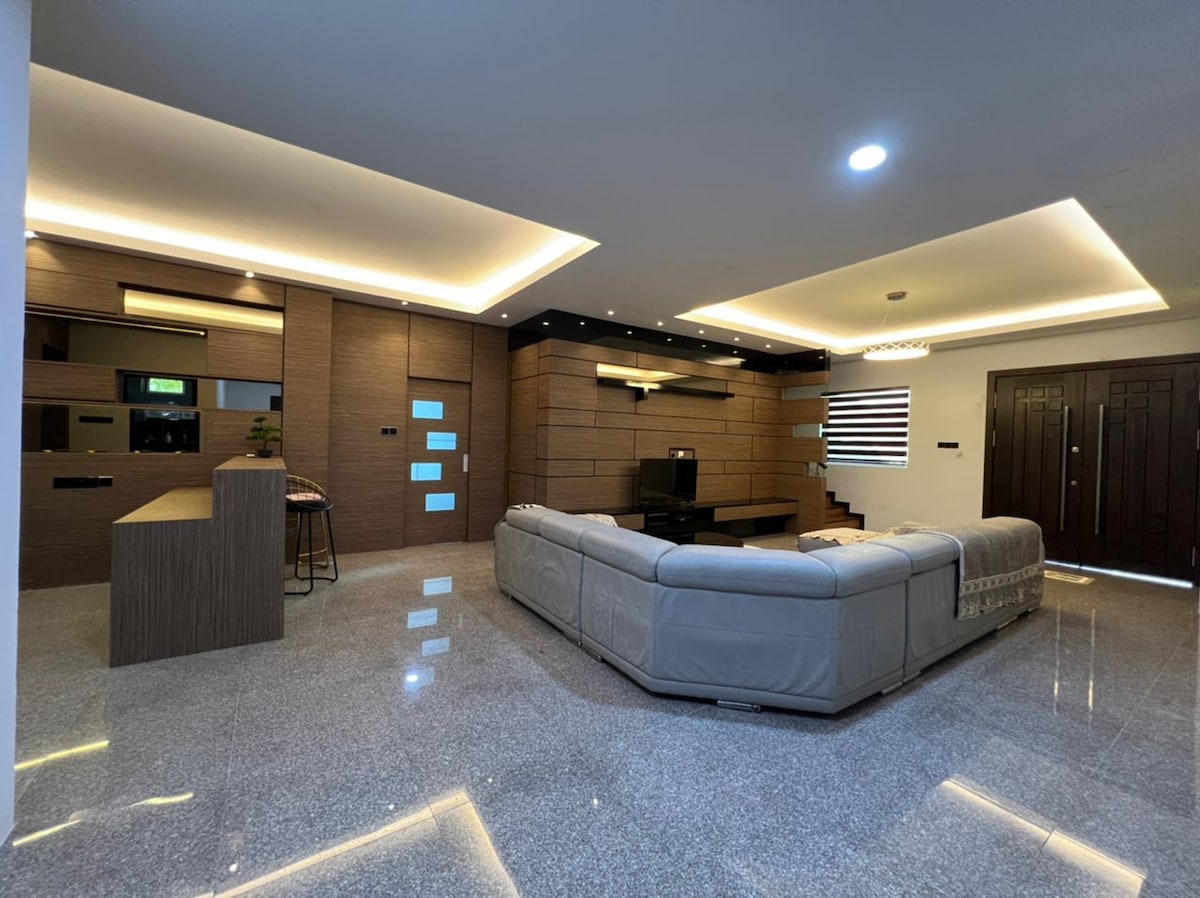A modern living area features a spacious sectional sofa arranged around a low coffee table. Walls of warm wood paneling contrast with glossy tile flooring, while recessed lighting enhances the sophisticated design. Large entrance doors and windows provide ample natural light.