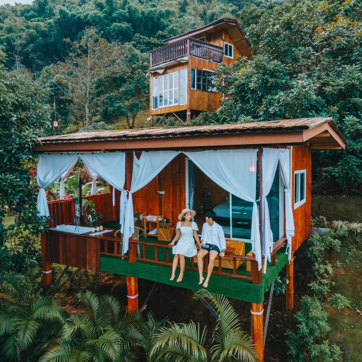 A wooden cabin is positioned on elevated posts, surrounded by lush greenery. Spacious openings allow natural light to fill the interior. A cozy outdoor seating area, under draped curtains, invites relaxation while providing views of the tranquil landscape.