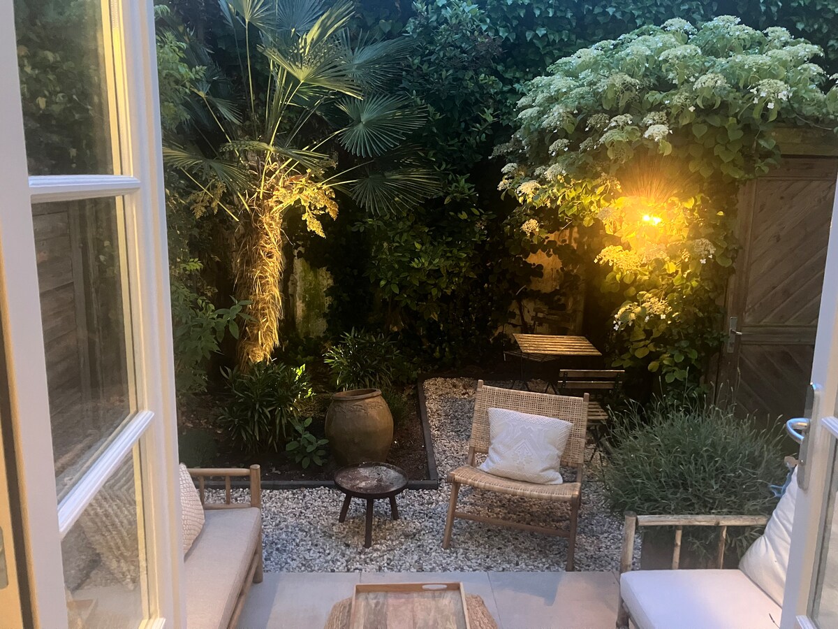 A tropical garden is featured, surrounded by lush green foliage. Soft lighting illuminates a cozy seating area that includes a small table and two chairs. A gravel path leads through the garden, enhancing the serene outdoor atmosphere.