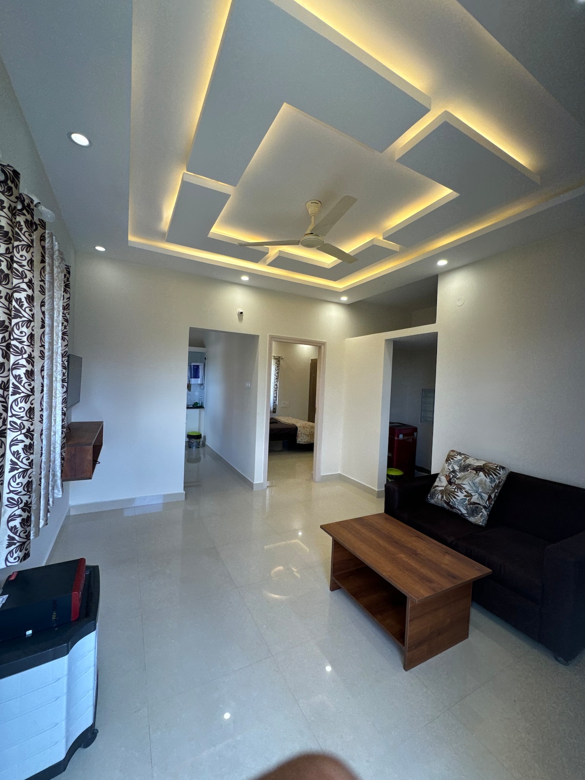 A spacious living area features a dark sofa and a wooden coffee table. Soft lighting highlights a modern ceiling design. Large windows with curtains allow natural light to fill the space, creating a bright and airy atmosphere. A glimpse of additional rooms can be seen in the background.