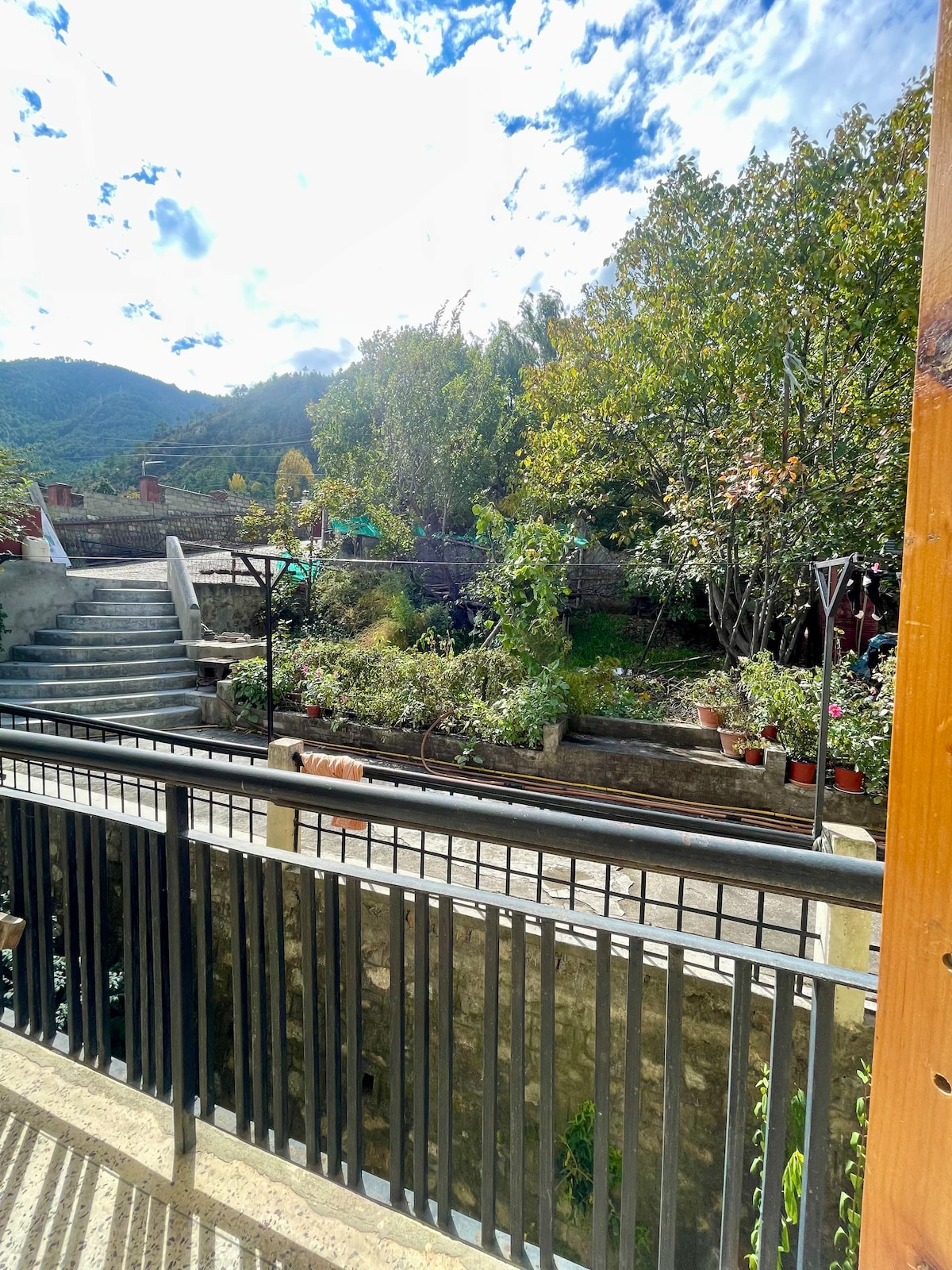 A serene garden area is visible, featuring a variety of lush green plants and trees. A stone pathway leads through the garden with stairs on one side. A peaceful mountainous backdrop enhances the natural setting under a partly cloudy sky.