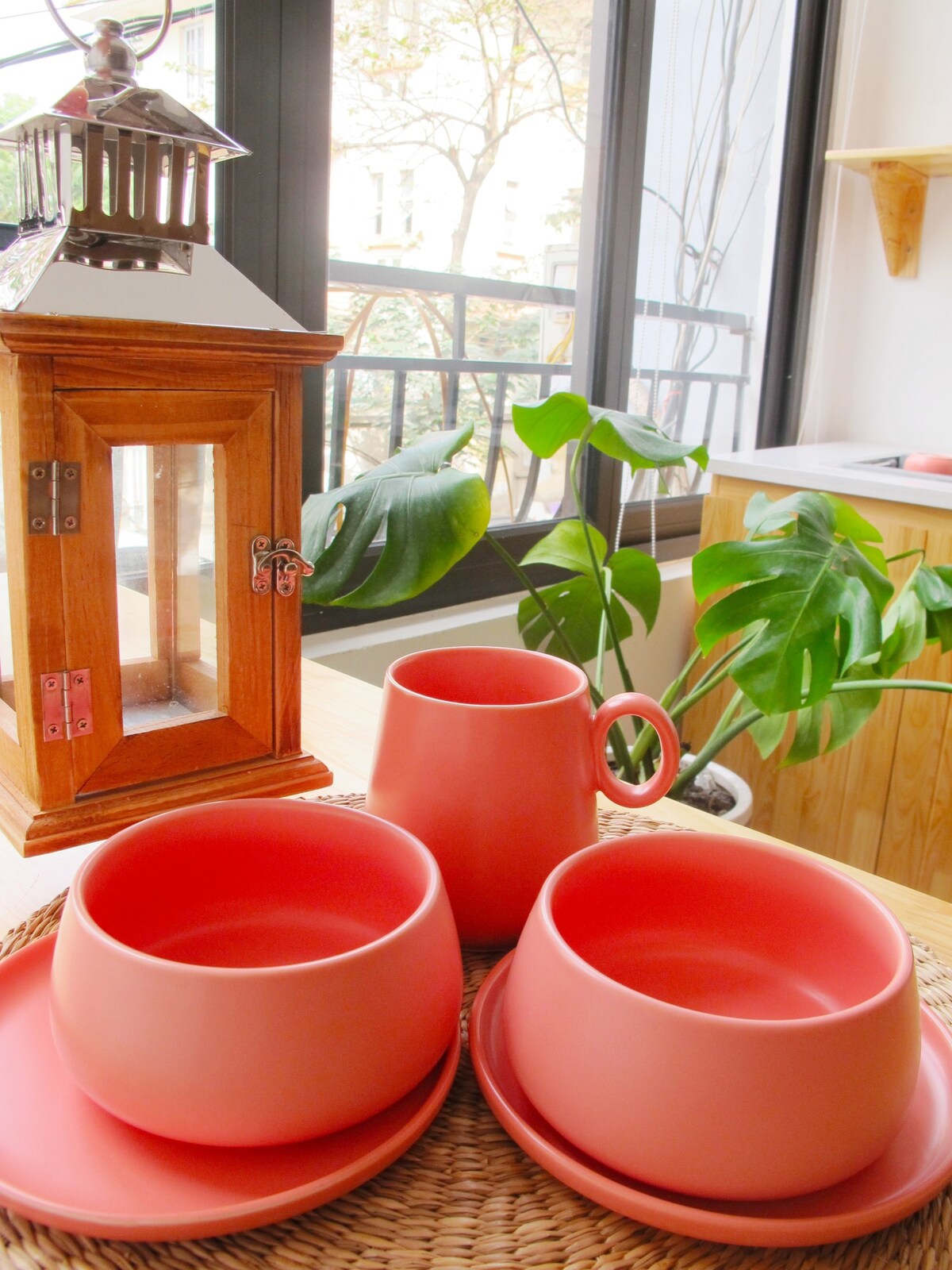 A wooden lantern stands next to a small plant, with vibrant coral-colored tableware arranged on a woven mat. Two bowls, a plate, and a cup showcase a modern design, complemented by natural lighting filtering through a large window.