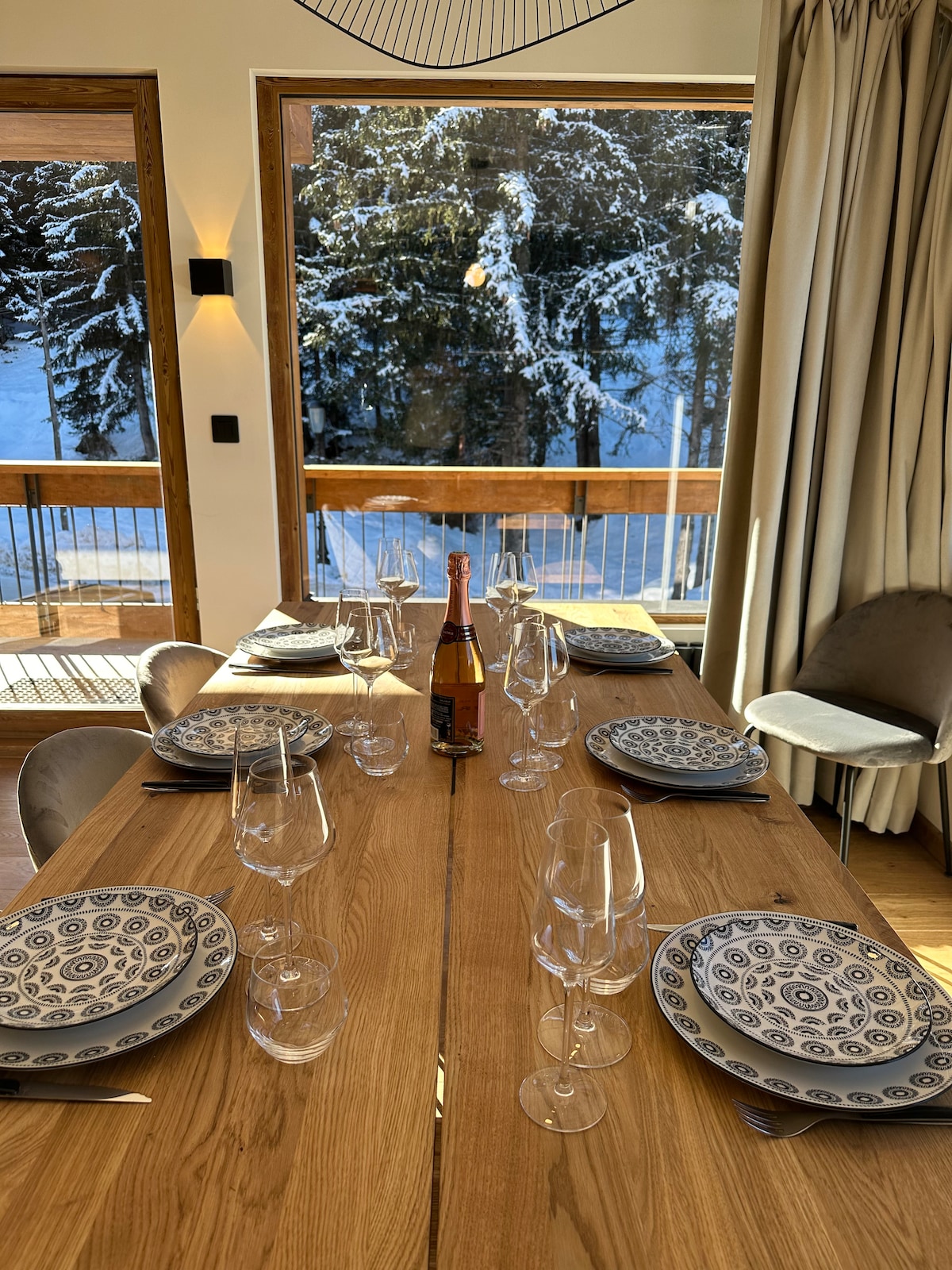 A dining table is set with elegant dinnerware, featuring patterned plates and clear glassware. A bottle of champagne is placed centrally. Large windows showcase a snowy landscape outside, while warm light enhances the wooden tones of the interior.