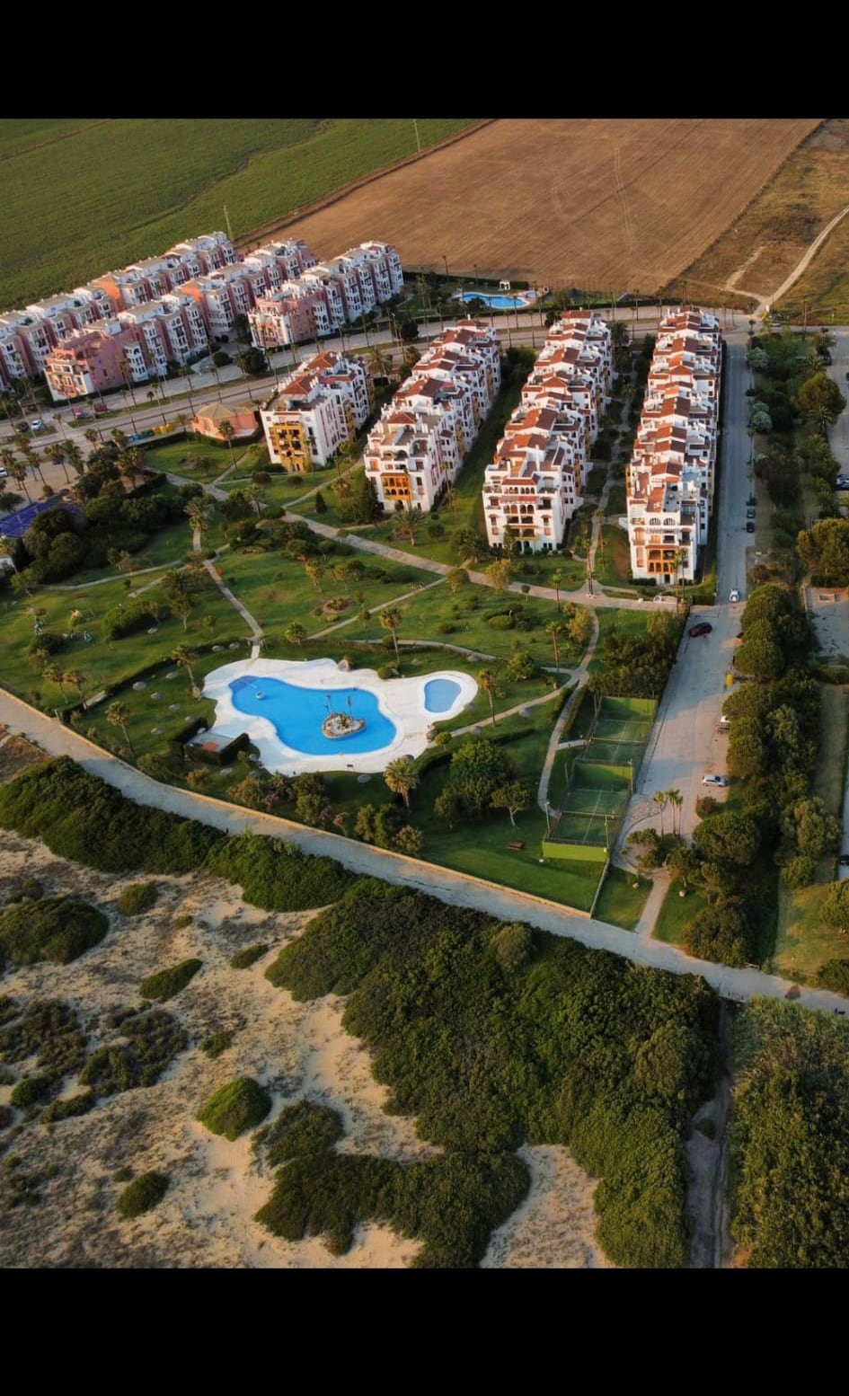 An aerial view captures a landscaped area featuring a large swimming pool surrounded by greenery. Nearby, residential buildings are visible with a mix of red rooftops and white facades, complemented by open green spaces and pathways interspersed throughout the area.