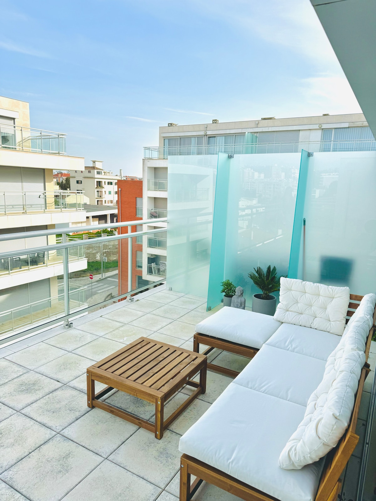 A spacious terrace features a wooden table and a large white cushioned seating area. Glass panels provide a sense of openness and protection while allowing natural light to fill the space. Potted plants are placed near the seating, creating a relaxing outdoor atmosphere.