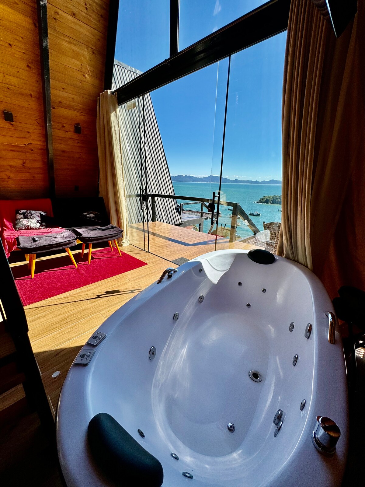 A spacious hot tub is placed near a large glass wall, offering views of the outdoor landscape. A seating area with two chairs and a small table can be seen, along with a red rug on the wooden floor, enhancing the inviting setting.