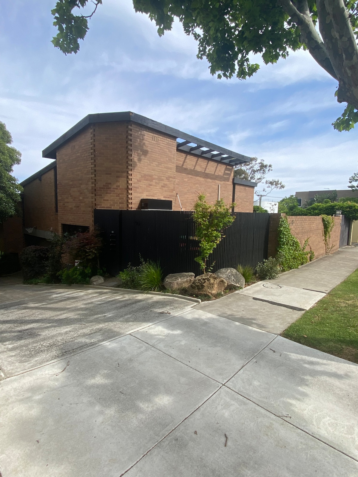 A contemporary brick building is partially obscured by a tall black fence, creating a private atmosphere. The structure features a sloped roof and is surrounded by greenery, including trees and shrubs. A wide concrete pathway leads to the entrance, with a well-maintained lawn beside it.
