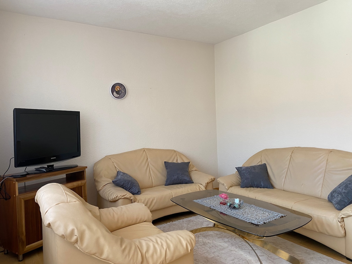 A light and neutral living area features a glass coffee table positioned between two beige sofas with decorative blue cushions. A wooden entertainment unit holds a television, and a subtle wall clock adds to the decor. Natural light fills the space, enhancing its openness.