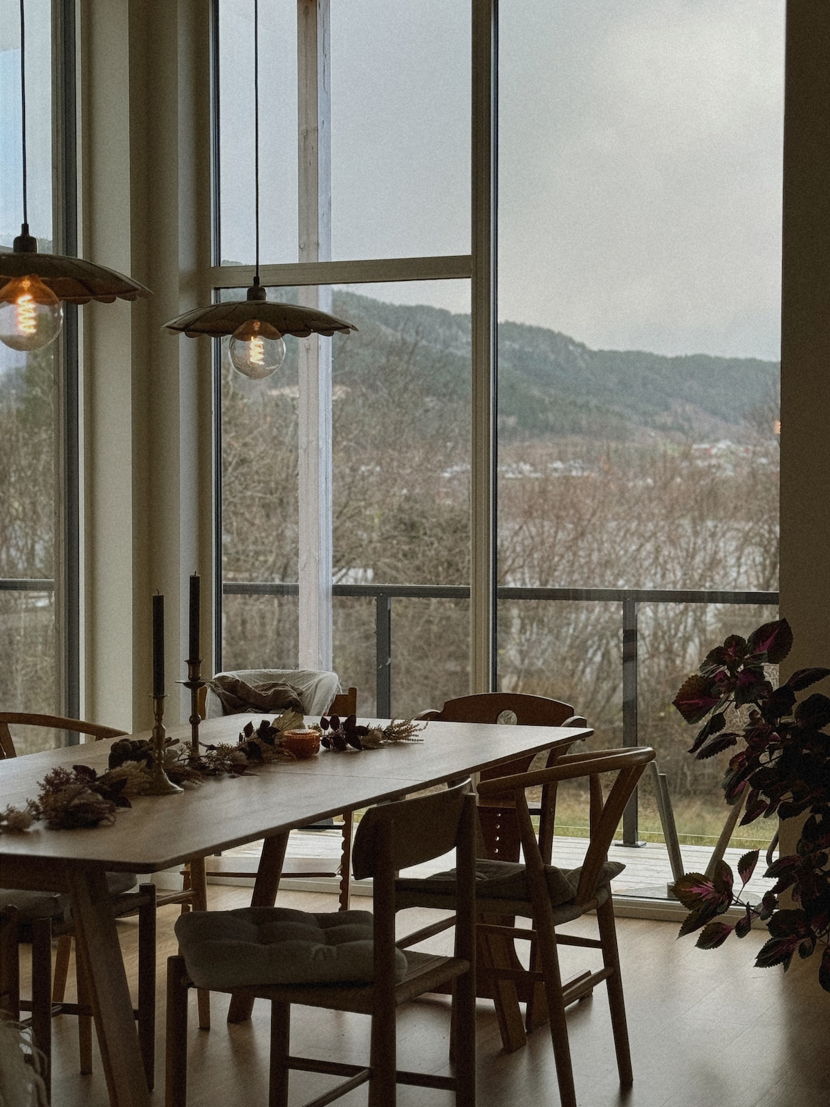 A dining area is set with a wooden table and mismatched chairs, surrounded by large windows that frame views of mountains and trees. Pendant lights provide soft illumination, and a decorative centerpiece adds a touch of elegance to the space.