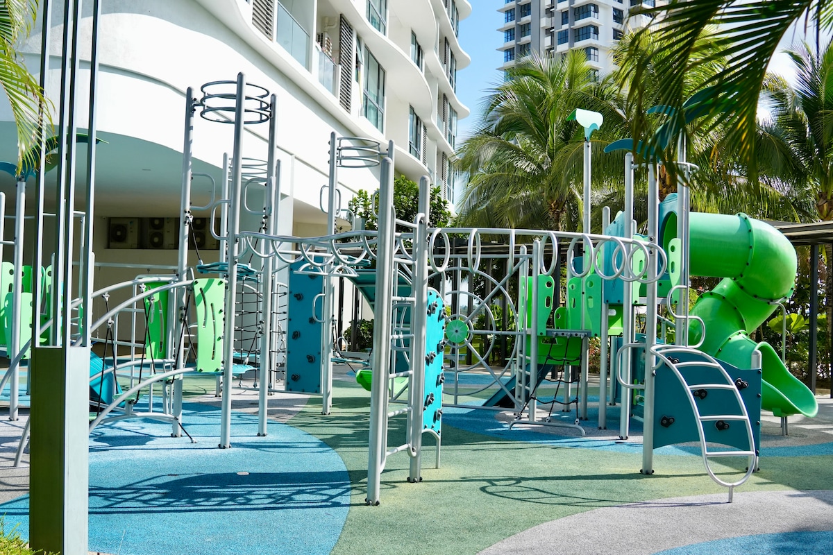 An outdoor play area is depicted, featuring a variety of green and blue equipment, including climbing structures, slides, and an obstacle course. Surrounding palm trees add to the environment, creating a space designed for recreational activities.