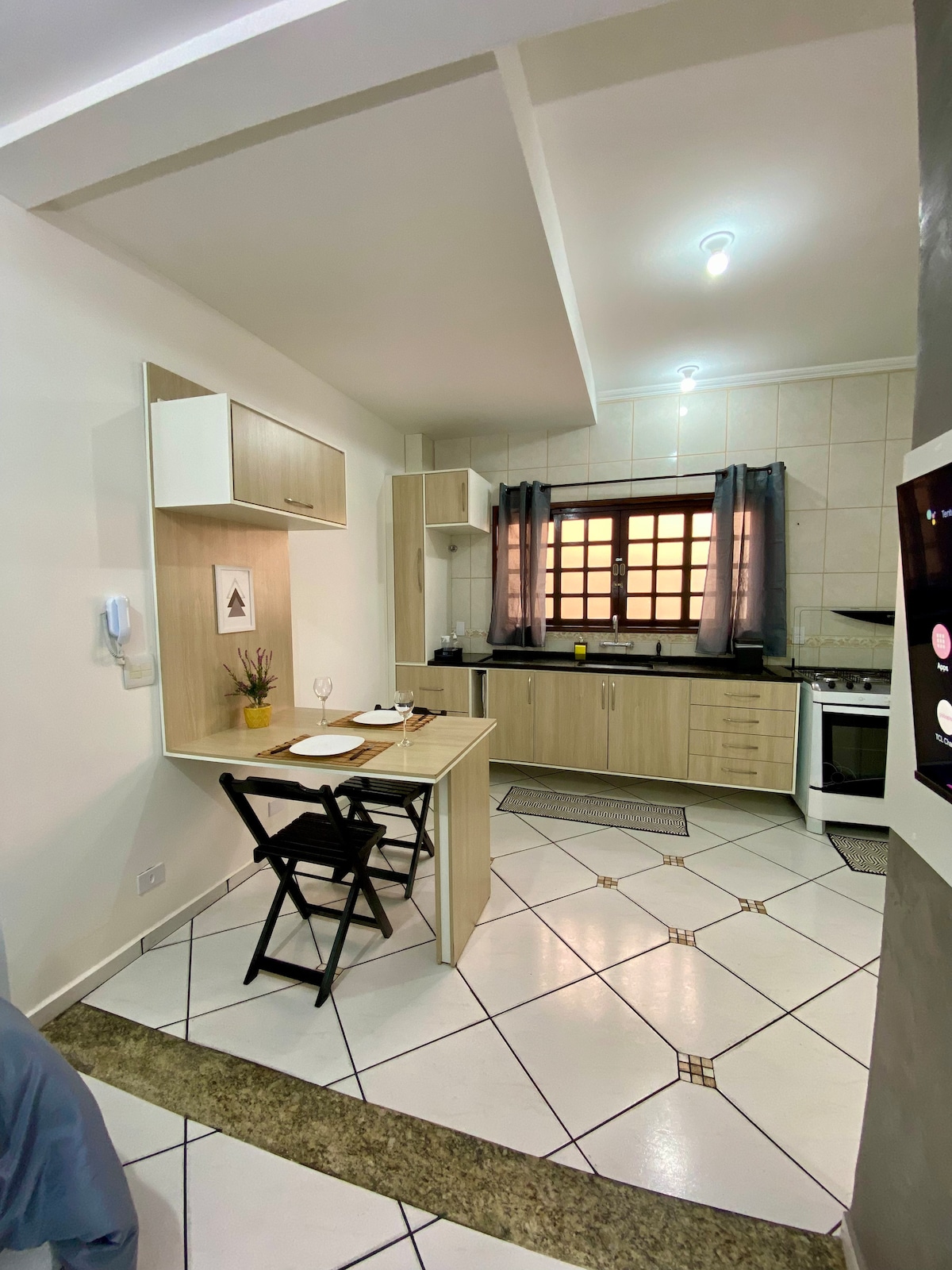 A modern kitchen area features a compact dining table with two chairs, positioned near a window dressed with gray curtains. The kitchen counter displays essential appliances, while light tiles create a bright atmosphere. A subtle decorative element adds a touch of style to the space.