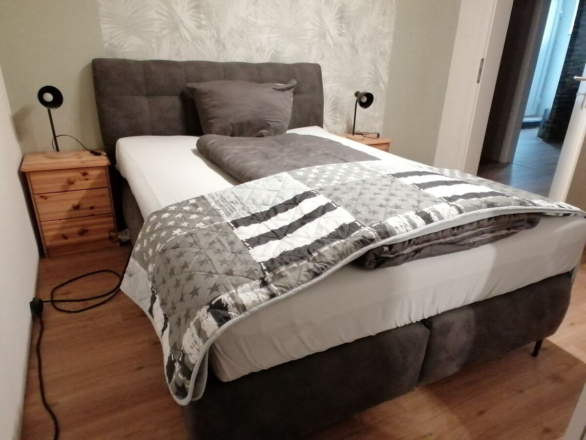 A spacious bed is positioned against a neutral wall with a subtle leaf pattern. Two bedside lamps are located on wooden nightstands, providing functional lighting. The bed is adorned with a quilt featuring a star pattern, creating a cozy and inviting sleeping environment.