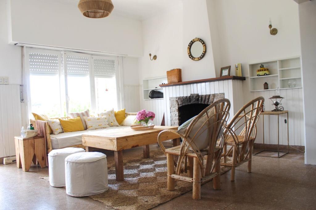 A light-filled living area features a comfortable sofa adorned with yellow and white cushions. A wooden coffee table sits in front of the sofa, complemented by two rattan chairs and a small round ottoman. A cozy fireplace is set against a streamlined white wall.