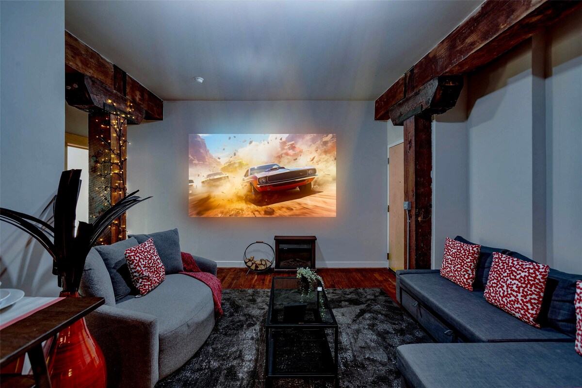 A spacious living area features a large projection screen displaying a vibrant movie scene. Two sofas are arranged for comfort, adorned with colorful cushions. A coffee table is centered on a plush area rug, with decorative plants adding a touch of warmth to the space.