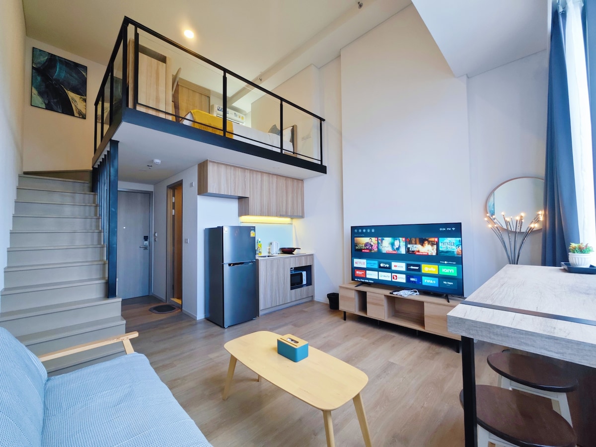 An open living area showcases a staircase leading to an upper level. A kitchenette is visible, equipped with modern appliances. A television is mounted on the wall, and a light-colored sofa is present alongside a wooden table. Natural light pours in through large windows, enhancing the space.