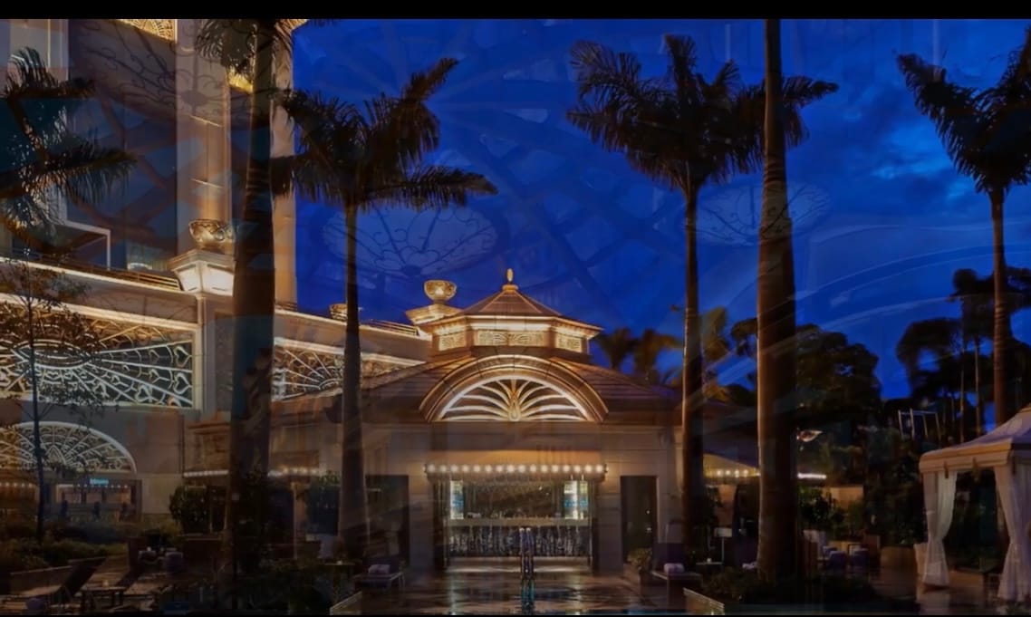 The exterior of the hotel is illuminated at dusk, showcasing a grand entrance. Palm trees frame the entrance, and architectural details are highlighted by soft lighting. Reflections from the glass facade accentuate the building's elegance against a deep blue sky.