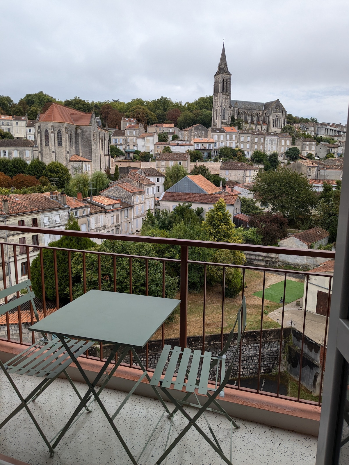 A small balcony is furnished with a table and two folding chairs. Below, a scenic view of Angoulême's plateau is displayed, featuring historic buildings and greenery, with a church steeple visible in the distance. The soft colors of the rooftops complement the natural landscape.