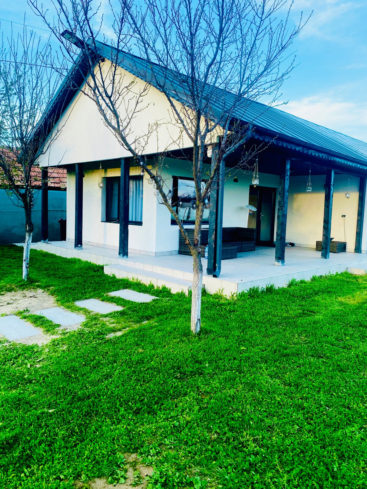 The exterior of Tacchini’s Home is shown, featuring a light-colored facade and a sloped roof. A well-maintained lawn surrounds the house, with stepping stones leading to a porch area. Leafless trees stand beside the home, and windows allow natural light to brighten the space.