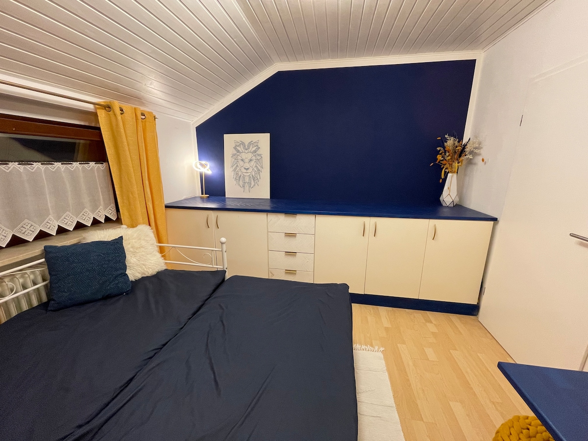 A cozy bedroom features a spacious bed with dark bedding and decorative pillows. A sleek blue accent wall complements light-colored furniture, including a dresser and a small table. Soft yellow curtains frame the window, adding a touch of warmth, while a decorative bouquet provides a subtle accent.