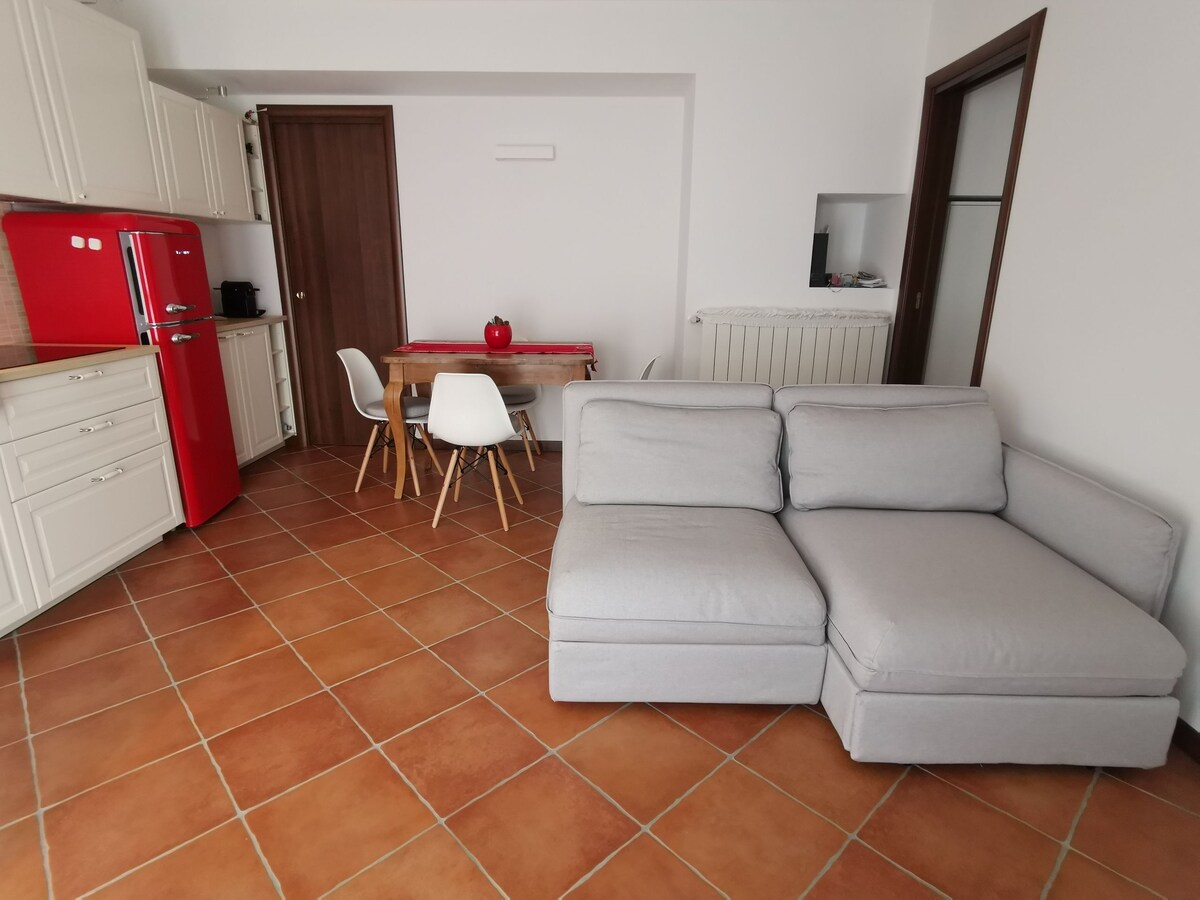 A modern kitchen is complemented by a vibrant red refrigerator, while a wooden dining table is surrounded by four chairs. A light gray sectional sofa is positioned in the living area, with tiled flooring adding to the inviting ambiance.