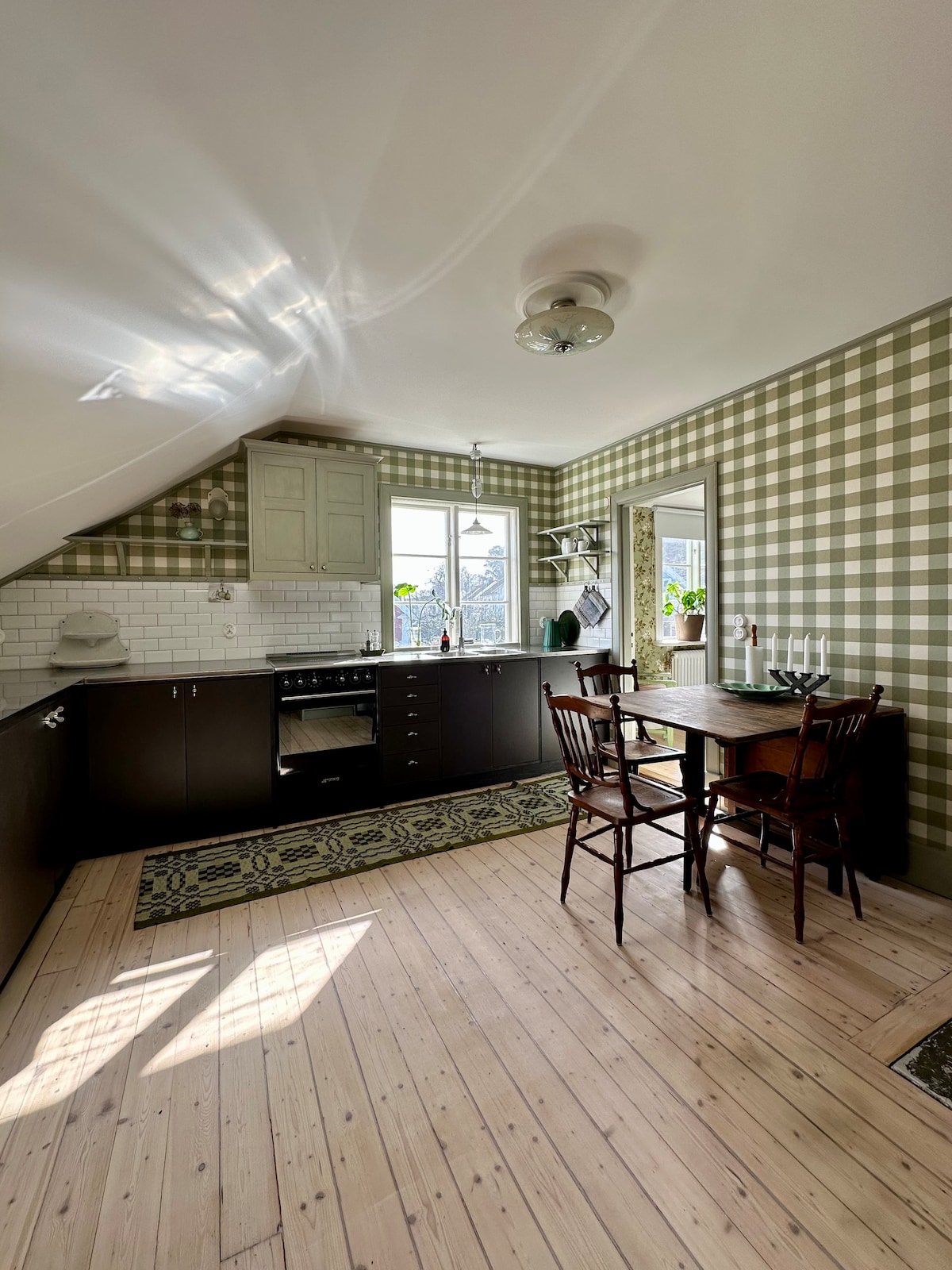 The kitchen features a modern design with plaid wallpaper and a light wood floor. Dark cabinetry contrasts with white tiles above the stove. A dining area is set with a small table and four wooden chairs, illuminated by natural light from the window.