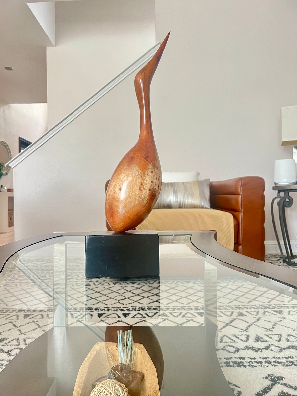 A wooden sculpture with an elongated, elegant form is positioned on a glass-topped coffee table. The piece stands out against a patterned rug. In the background, a leather couch and soft decorative pillows are visible, enhancing the modern living space.
