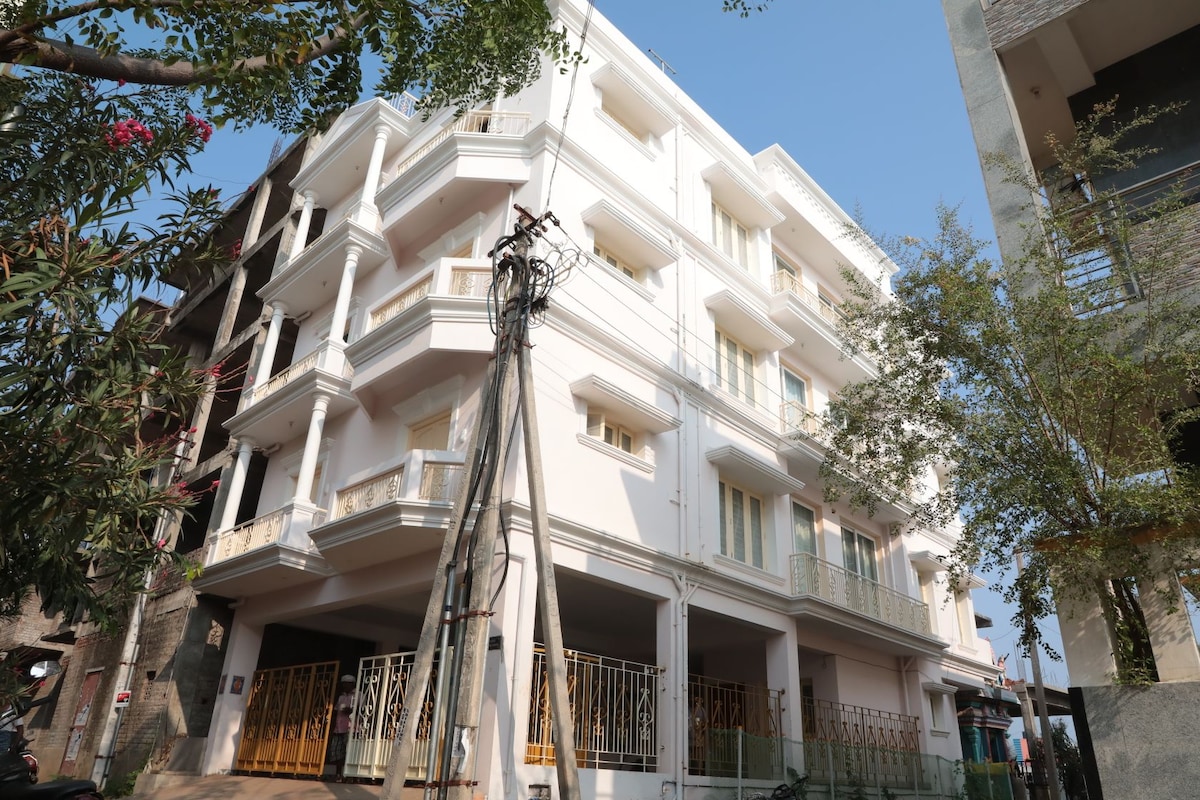 The modern apartment building features a clean, white facade with multiple balconies. Large windows allow ample natural light to flood the interior. A spacious entrance area with decorative metal gates is visible, along with greenery along the sides of the property.