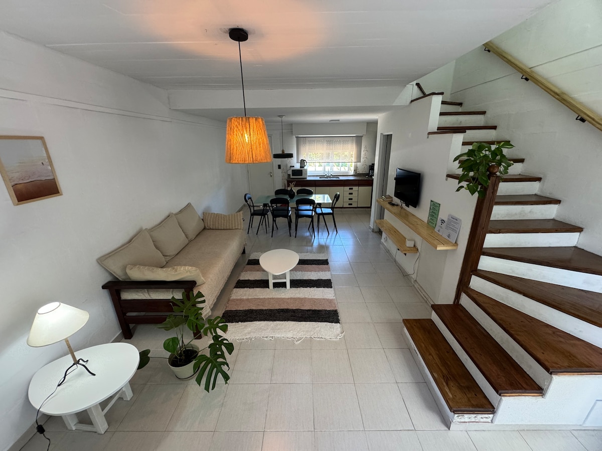The open living area features a light-toned sofa, a round coffee table, and a small floor lamp. Dining chairs are arranged around a table in the kitchen space, which includes modern appliances. A staircase leads to the upper level, enhancing the spacious layout.
