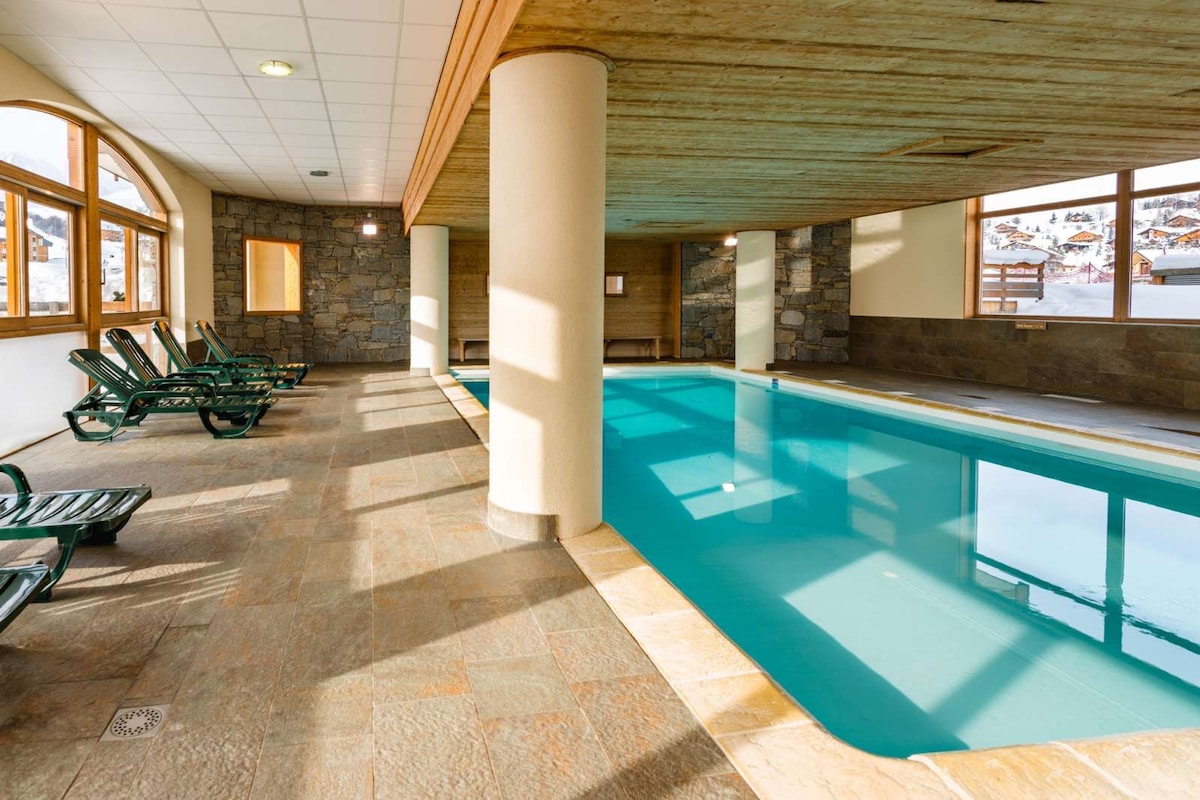 An indoor swimming pool is surrounded by natural stone walls and large windows that allow for ample natural light. Lounge chairs are placed beside the pool, providing a relaxing space for guests. A wooden ceiling adds warmth to the modern design.