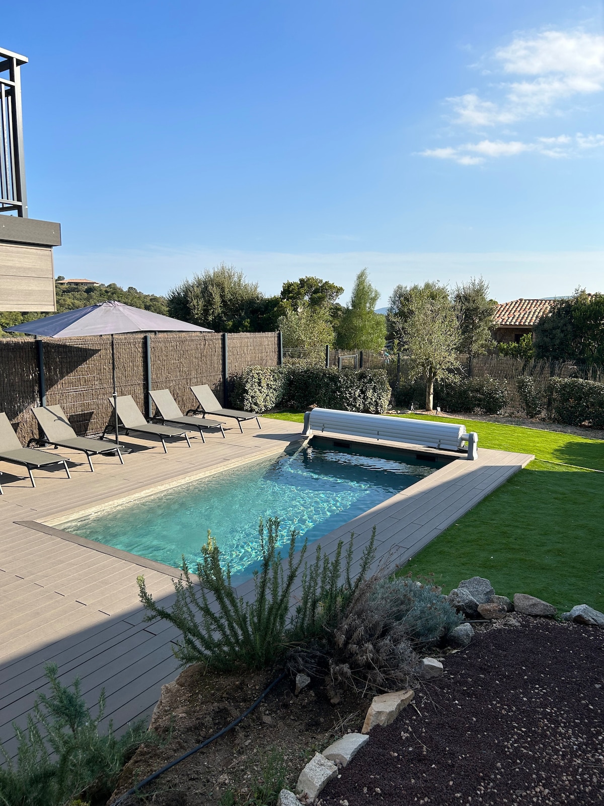 A tranquil outdoor space features a private salt-water pool surrounded by a wooden deck. Sun loungers are arranged nearby, and a shaded area under a canopy offers respite. Lush greenery and shrubs provide a natural backdrop, enhancing the inviting atmosphere.