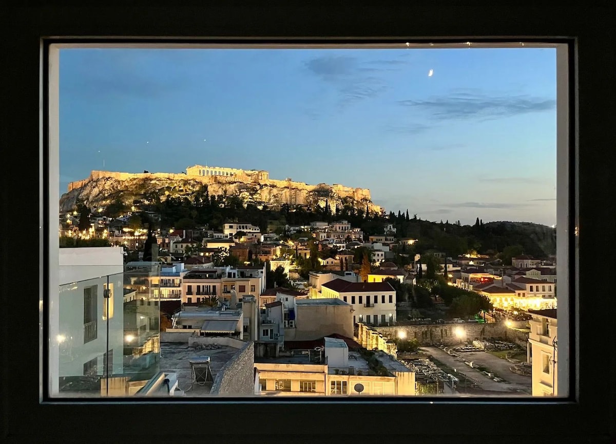 A panoramic view from a window features the Acropolis illuminated at dusk, surrounded by the city of Athens. The scene captures a blend of traditional architecture and modern buildings, with soft lighting creating a serene atmosphere as the sky transitions from blue to twilight.