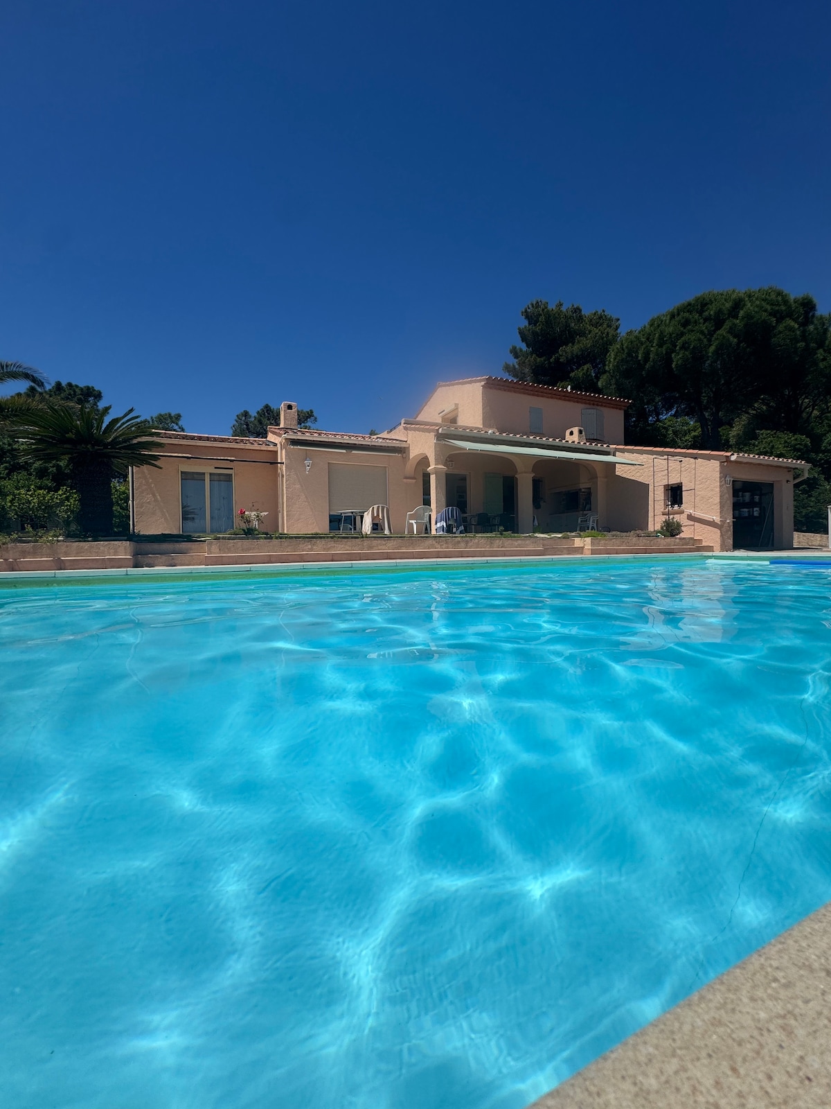 The villa is viewed from the pool, showcasing a sunlit exterior surrounded by greenery. Clear water reflects the sky, enhancing the inviting atmosphere. A terrace with seating is visible, while palm trees add to the tranquil environment.