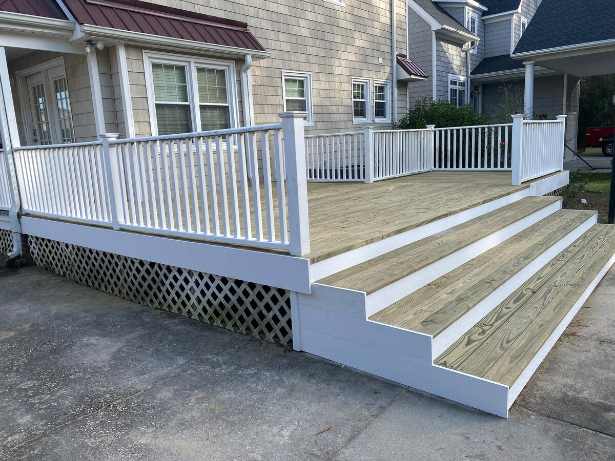 A spacious deck with white railings and steps is visible, leading up from a concrete area. The wooden flooring of the deck provides a warm contrast to the white structure. Several windows from the house are seen in the background, indicating a welcoming outdoor space.