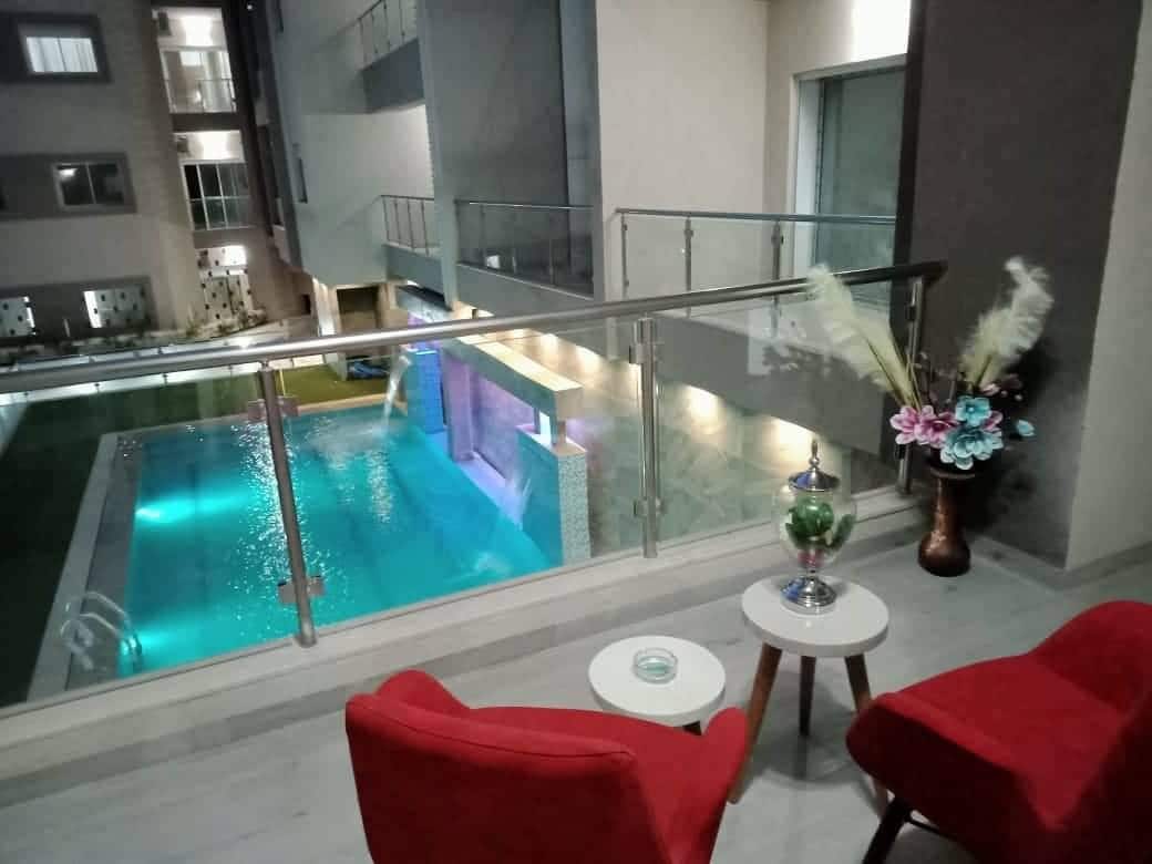 A balcony view showcases a luminous swimming pool surrounded by soft lighting, with modern architecture visible in the background. Two red armchairs are positioned beside a small table, creating an inviting area for relaxation.