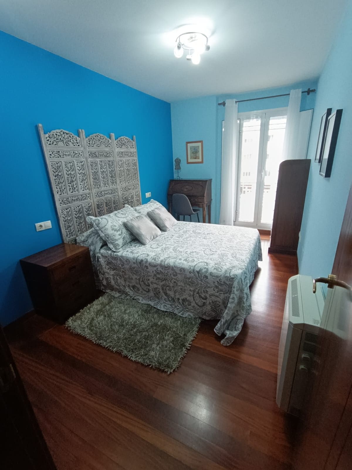 A serene bedroom is presented with a vibrant blue wall. A spacious bed is adorned with patterned bedding, accompanied by two nightstands. A decorative screen adds a unique touch, while a desk and chair are positioned near a window that provides natural light.