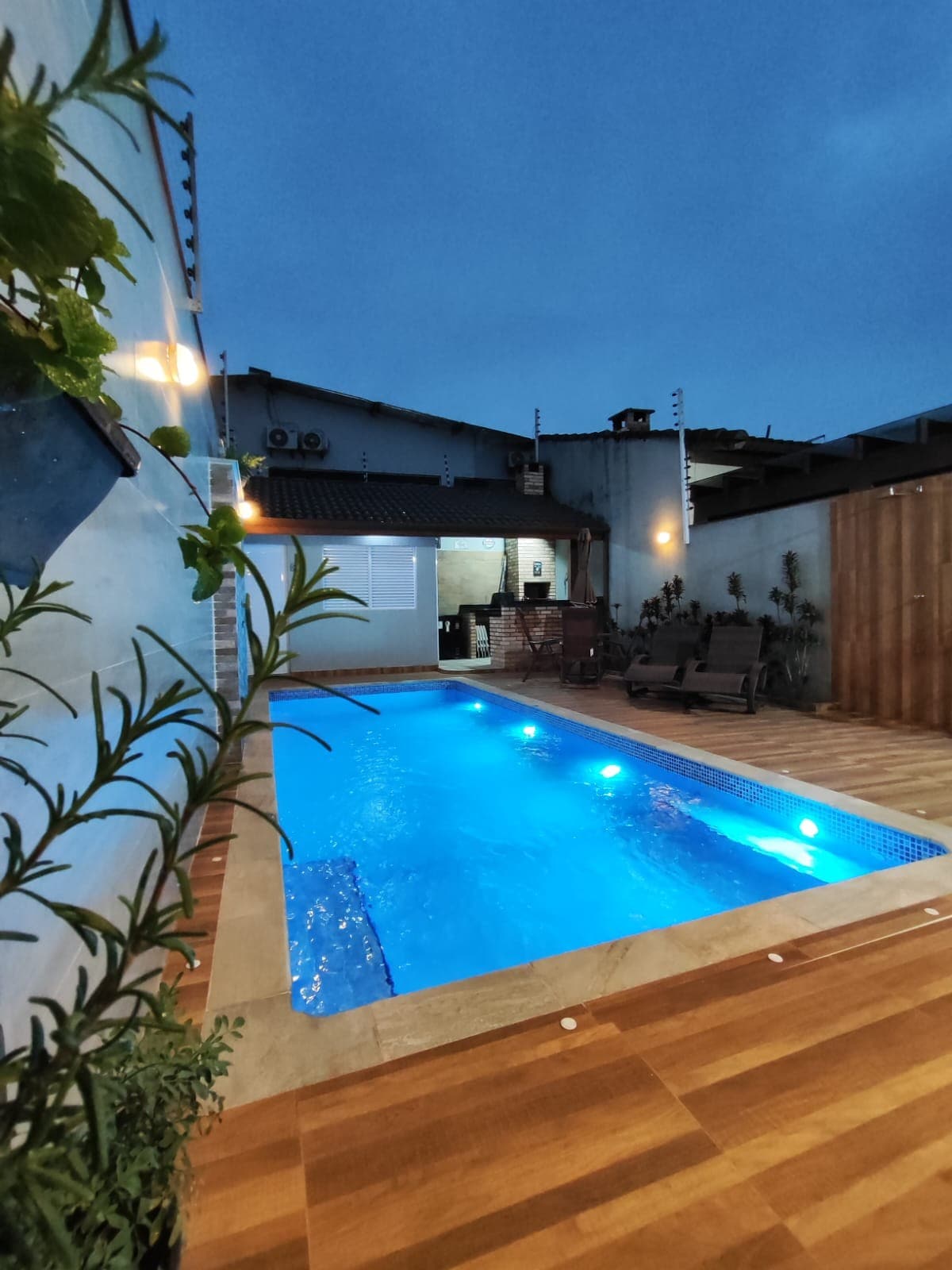 An inviting outdoor space features a well-lit pool with calming blue water, surrounded by wooden decking. Plant life adds a touch of greenery, while a seating area with benches can be seen in the background, enhancing relaxation options for guests.
