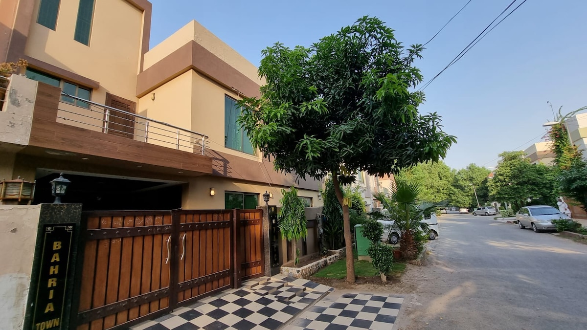 A modern exterior is showcased with a double-gated entrance and a checkered pathway leading to the entrance. Lush greenery surrounds the property, and a large tree provides shade, while the peaceful street features a few parked cars.