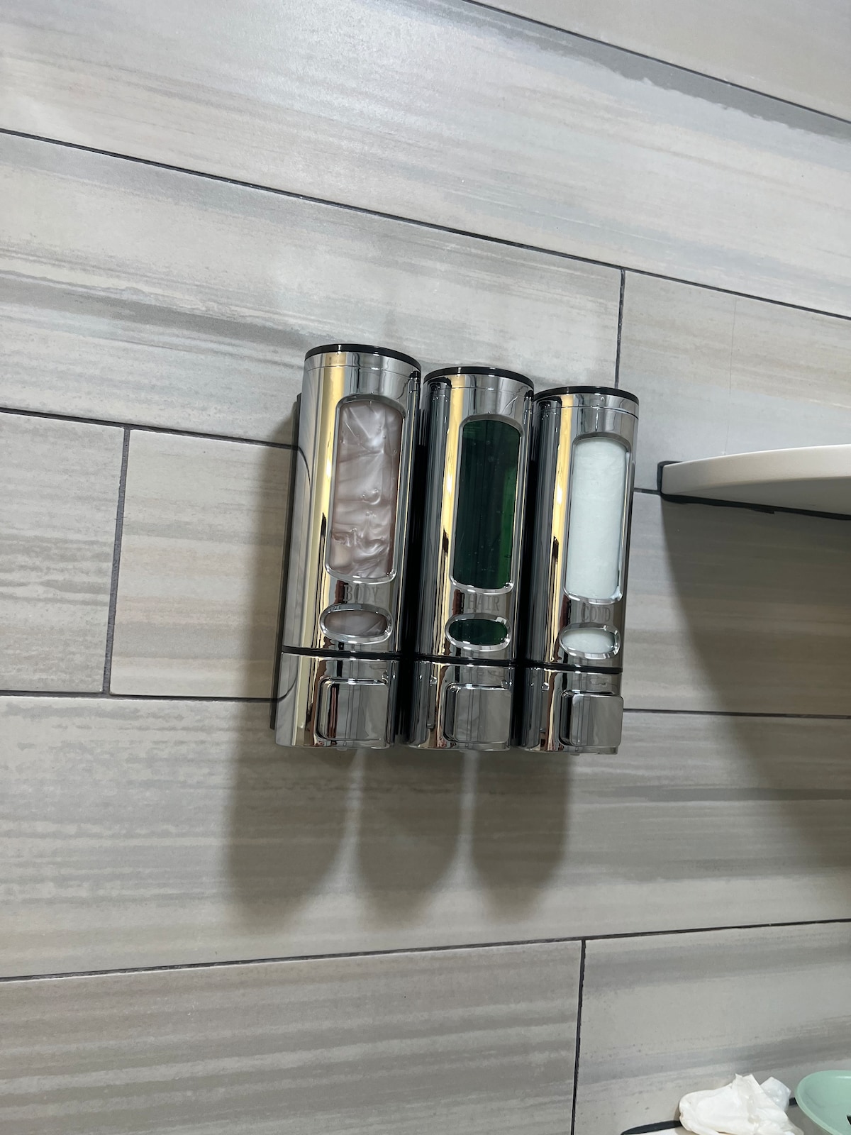 Three chrome wall-mounted dispensers are arranged in a row against a backdrop of light grey, textured tiles. Each dispenser contains complimentary liquid products, clearly labeled for easy identification, enhancing the bathroom's functionality and modern aesthetic.