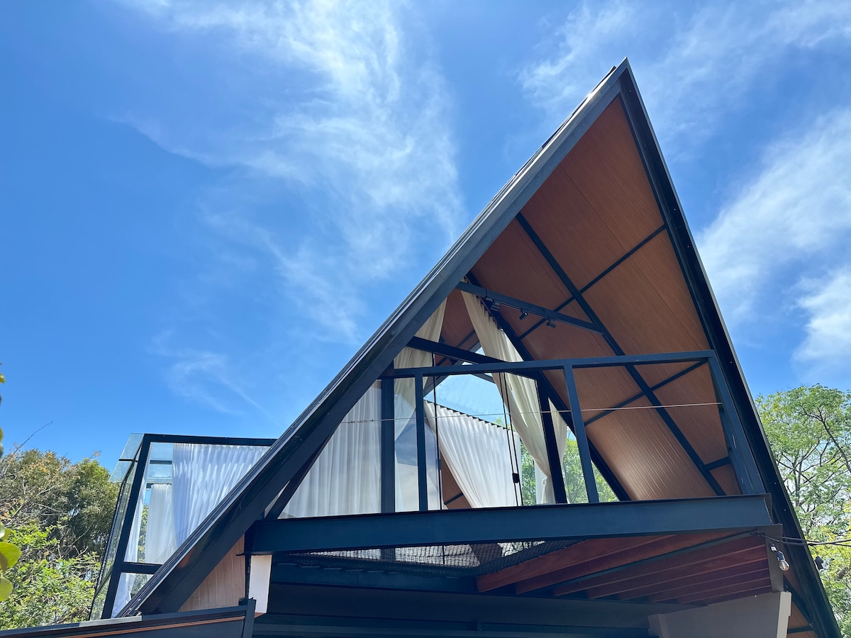 A modern triangular roof structure is visible against a bright blue sky, featuring a combination of wood and metal materials. Large glass panels are integrated into the design, allowing for natural light and scenic views of the surrounding greenery.
