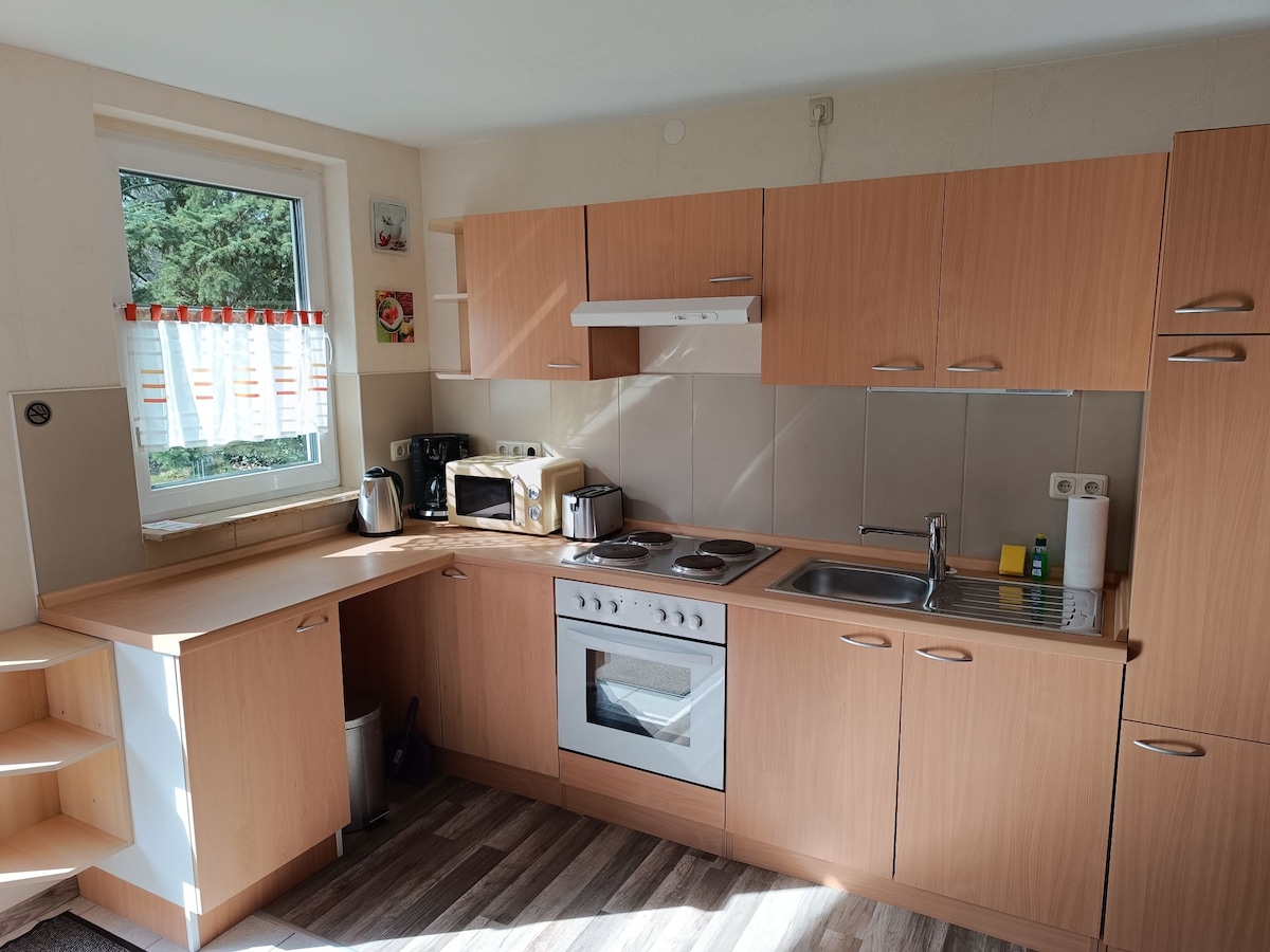 The kitchen features light wood cabinetry and a spacious countertop. Essential appliances include a stove, oven, microwave, and kettle, with a sink conveniently located. A window with sheer curtains allows natural light to illuminate the area, creating a bright and functional kitchen space.