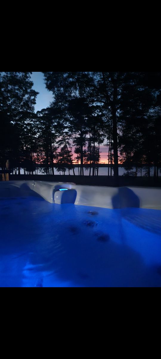 A calming hot tub is visible, reflecting soft blue lights, positioned near a serene lakefront. Tall trees frame the scene, with a colorful sunset visible in the background, highlighting the tranquil atmosphere of the outdoor space.