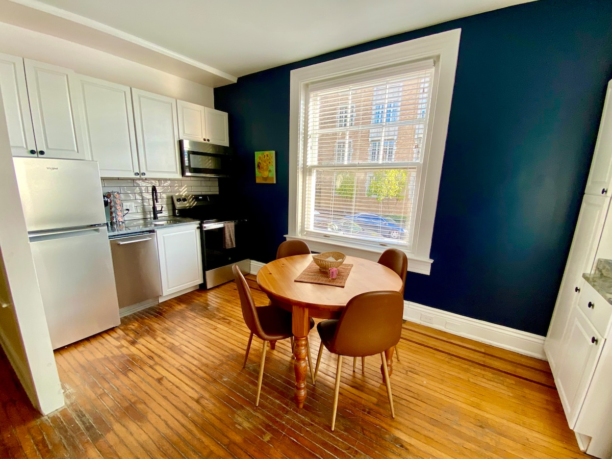 A cozy eat-in kitchen features modern appliances, including a stainless-steel refrigerator and an oven. A round dining table with four brown chairs is positioned near a large window, allowing natural light to brighten the space. Exposed wooden flooring adds warmth and character.