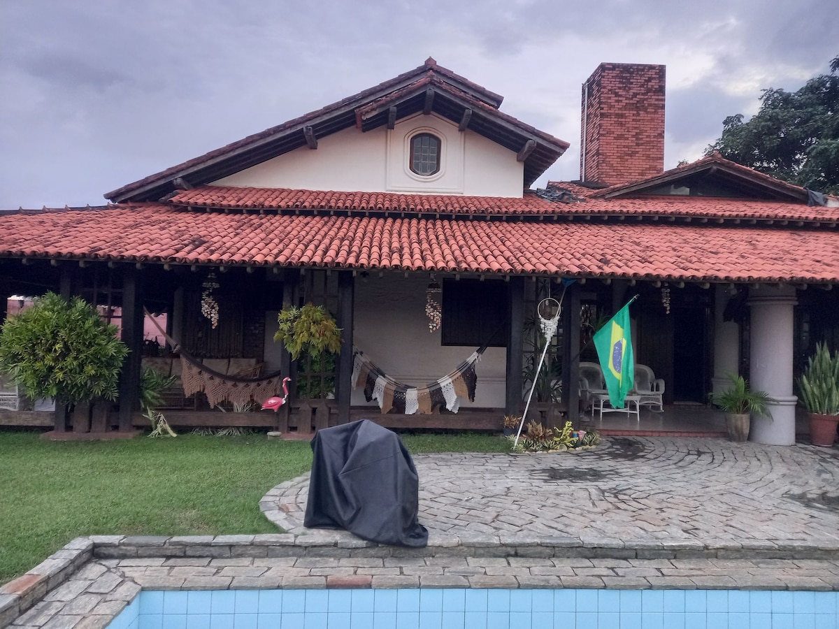 A spacious house with a traditional design features a terracotta tiled roof and a covered outdoor area. A Brazilian flag flutters near neatly arranged plants. A charcoal grill is covered, and a gentle lawn extends around the swimming pool in the foreground.