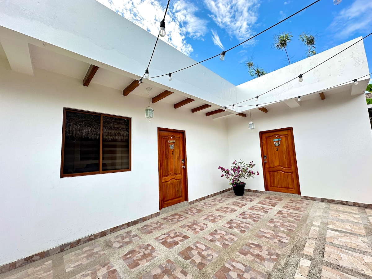 An outdoor area features two wooden doors with decorative handles, flanked by a potted plant. The ground is paved with stone tiles arranged in a patterned layout, while strings of lights are suspended above, adding a subtle charm to the space.
