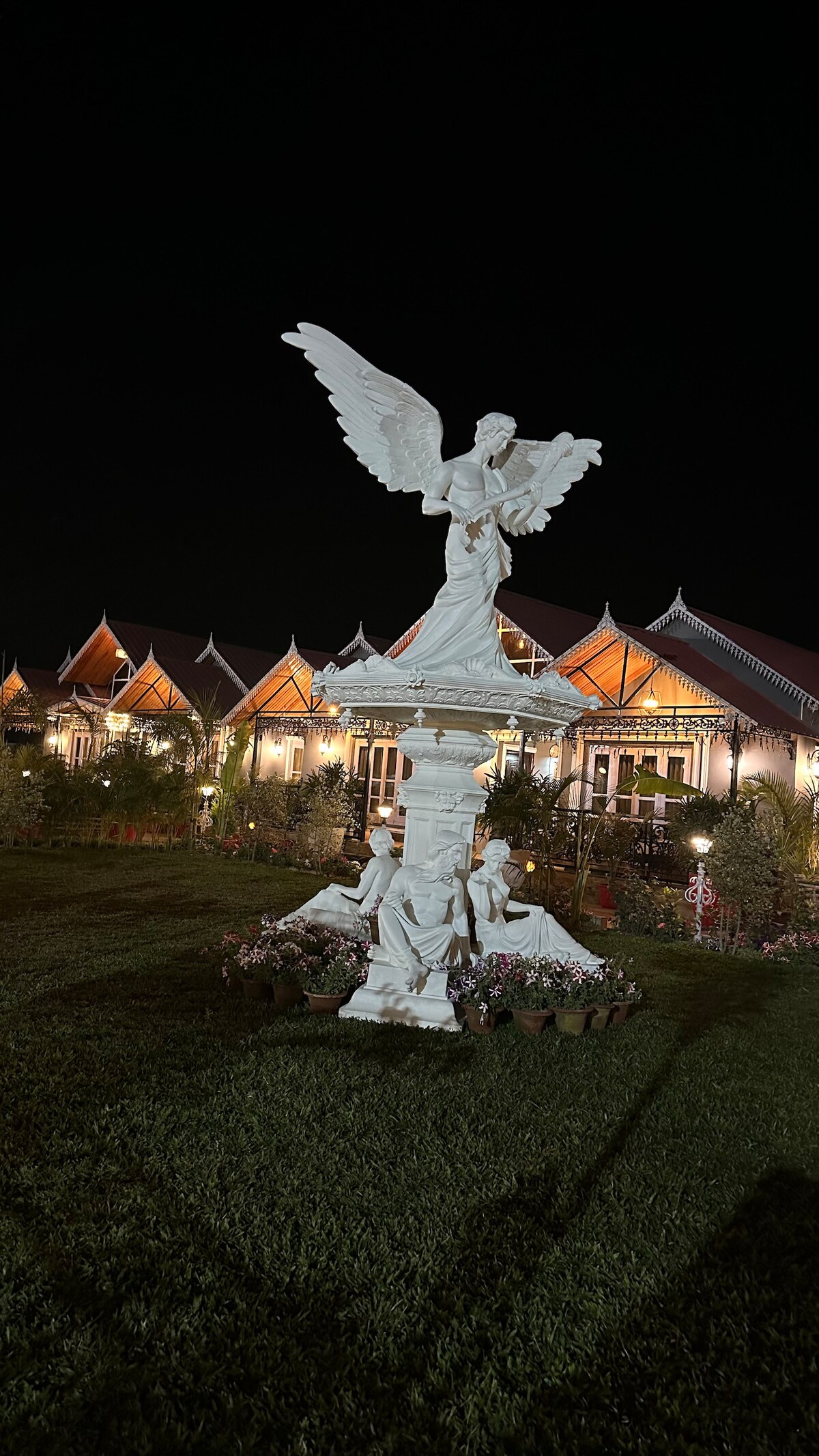 A striking white statue of an angel with outstretched wings is positioned in a well-kept garden. The background features a charming villa illuminated against the night sky, surrounded by lush greenery and decorative plants.